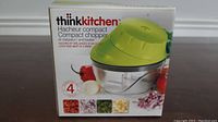Front view of the boxed thinkkitchen compact chopper showing green lid and clear container with tomatoes and onion around it.