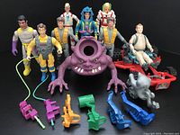 Photo showing 10 Ghostbusters figures including a large purple monster, one seated on a red vehicle, and multiple proton pack accessories on black background.