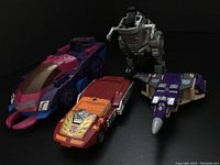 Four 1980s Transformers figures in vehicle robot modes on a dark surface: red and orange Hot Rod, purple Blitzwing, and Road Grabber, plus silver Grimlock dinosaur robot.