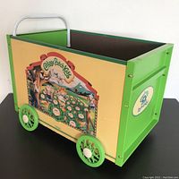 Side view of the 1984 Cabbage Patch Kids toy cart highlighting the colorful side panel artwork and green wheels, showing moderate surface wear and overall structure.