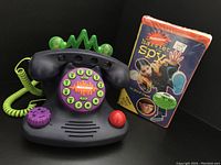 Photo showing the 1997 Nickelodeon Talk Blaster phone and sealed 1996 Harriet The Spy VHS movie from an angle highlighting the phone's dial and VHS cover