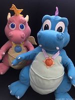 Photo showing both Cassie and Ord Dragon Tales plush dolls sitting against a black background. Cassie is pink with yellow accents and Ord is blue with a purple horn.