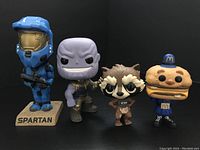 Four Funko POP! figures including Spartan, Thanos, Rocket Raccoon, and McDonald's hamburger character arranged side by side on a black surface.