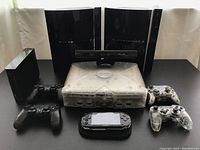 Photo showing two Sony PlayStation 3 consoles, one Xbox console with Kinect sensor, transparent Nintendo Wii console, multiple controllers and PSP handheld device.