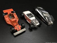 Three Hot Wheels cars on black surface showing front and side views. Orange Land Lord, white Ford Roadster with flames, and silver Shadow Jet II.