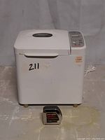 Front view of white Breadman breadmaker with digital control panel and top lid viewing window. Marked with '211' on front.