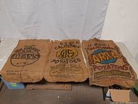 Three burlap bags laid out horizontally on the floor, showing front sides with agricultural branding for beans and potatoes.