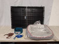 Photo showing black foldable metal pet cage, gray dog bed in plastic, blue heated pet bowl, and two dog collars/leashes (brown and blue) placed on a flat surface.