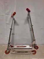 Two Razor kick scooters side by side, showing orange grips and wheels, silver decks with Razor logos