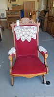 Front view of the antique Eastlake carved wooden platform rocking chair with red fabric upholstery and white crocheted doilies on headrest and armrests.