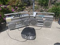 Front view of two vintage Panasonic boom boxes and one black Panasonic CD radio placed outdoors on concrete, surrounded by plants.