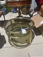Multiple angled photos of a round brass frame side table with tinted glass top and bottom shelf. Table is seen outdoors on concrete with other furniture in background.