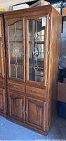 Full view of the oak wood curio cabinet showing two glass doors with decorative stained glass, two drawers beneath, and two wooden doors at bottom. Condition shows scratches and the right glass door is broken.