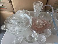 Glass decor items arranged on a surface, including cake stand, creamer and sugar set, trinket dishes, candle holder, vase, and basket.