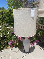 Front view of Mid-Century Modern lamp showing tall cream fabric shade and white ceramic base outdoors with flowering plants in background.
