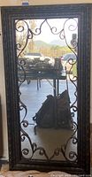 Full-length rectangular mirror with dark brown ornate scrolling metal frame, reflecting outdoor scene.