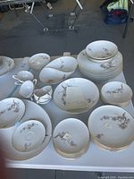 View of the full set displaying plates, bowls, cups, and serving dish arranged on a table. Items have a white base color with botanical branch pattern and gold rim accent.