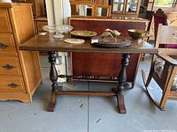 Front view of the vintage wood entryway table placed on a gray floor among other furniture. The table surface has visible scratches and decorative items on top which are not included in the lot.