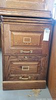Front view of a wooden 2-drawer filing cabinet showing brass handles, label holders, and keyhole on the upper drawer. Cabinet shows wood grain and minor surface wear at base.
