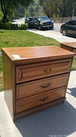 Front and side view of the wooden three drawer nightstand placed outdoors, showing drawer handles and some surface scratches.
