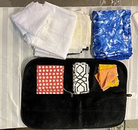 Top view of mixed fabric lot showing folded white gauze curtains folded on left, blue polyester blanket on right, black pet mat on bottom center with three sets of table napkins stacked on top.