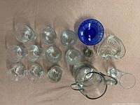 Glassware items arranged on cloth including pitcher, various glasses, blue dish, heart-shaped bowl, and shot glass.