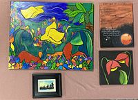 Four artworks displayed on a beige surface: large colorful acrylic painting with green, yellow, orange and purple, a small framed print, a watercolor painting with text and an image of a moon/sun, and a flower painting with red and pink on black background.