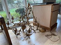 Full view of the gold metal chandelier on floor showing twelve arms with candle-style lights and decorative leaf motifs, along with hanging chain and ceiling canopy.