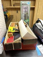 Box with assorted items including thermos box, wooden brush set box, notebooks, and metal emblems on basement shelf