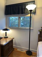 Photo showing the tall floor lamp with white shade illuminated next to a wooden nightstand with the black dome-shaped touch table lamp turned on.