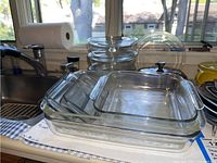 Four lidded Pyrex glass bowls stacked with visible knob handles on lids and three clear rectangular glass baking dishes stacked beside them.