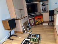 Two loudspeakers on stands in a room with wood floor and framed pictures on the floor.