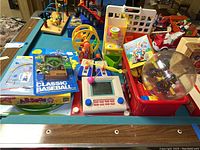 Overview of assorted vintage toys including boxed games, toy kitchen, electronic handhelds, and building blocks on a table.