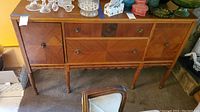 Front view of vintage buffet cabinet showing diamond-pattern veneer, drawer and door configuration, metal handles, legs.