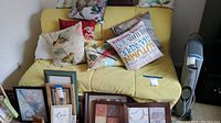 Folding futon day bed in yellow fabric cover shown folded up with multiple decorative throw pillows and framed pictures placed in front.