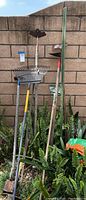 Wide shot showing all garden tools including rake, hoes, and metal stakes leaning against a brick wall above ferns.