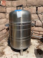 Front view of Meco metal outdoor charcoal grill showing cylindrical stainless steel body, domed lid with handle, and three metal legs.