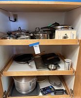 Three shelves inside a wooden cabinet filled with assorted pots, pans, lids, utensils, and a spiral-bound recipe book for pressure cooking.