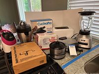 Lot on kitchen counter showing multiple small appliances including blender, toaster, crockpot, juicer, food processors, and boxed apple peeler and pizzelle baker.