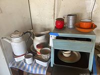 Photo of vintage enamel coffee pot, double-spout pitcher, small enamel cups, metal storage box, and other kitchen items arranged on a surface.