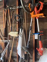 Wall-mounted yard tools including long-handled loppers, pruner, and hand sickle. Coiled extension cords hanging on hooks. Two Milwaukee cordless string trimmers visible with orange and black handles.