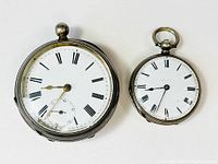 Both antique silver pocket watches shown side by side, highlighting their size difference and condition.