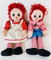 Front view of both Raggedy Anne and Andy dolls seated side by side showing clothing and facial details.