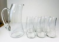Clear angled view of the glass lemonade pitcher and six matching glasses with bent curves arranged in a row, photographed on white surface against white background.