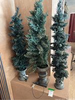 Three tall, narrow faux pine trees lit electrically, each in a stone-colored urn base. Shown indoors against a beige wall.