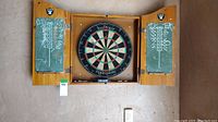 Full view of wooden dart board cabinet open, showing dartboard and chalk scoreboards with markings and Oakland Raiders stickers on cabinet doors.