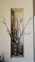 Full view of tall glass vase holding several faux cherry blossom branches with white flowers, placed in a niche on the wall.