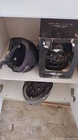 Two bike helmets and one hockey helmet inside a cabinet shelf. The hockey helmet is boxed, and the bike helmets show use.