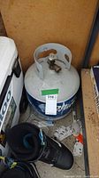 Blue Rhino propane tank with visible valve and metal handle, placed next to a cooler and snowboard boot.