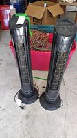 Two black vertical tower fans with cylindrical bodies and round bases, positioned on a concrete floor with a red plastic bin and other items in the background.